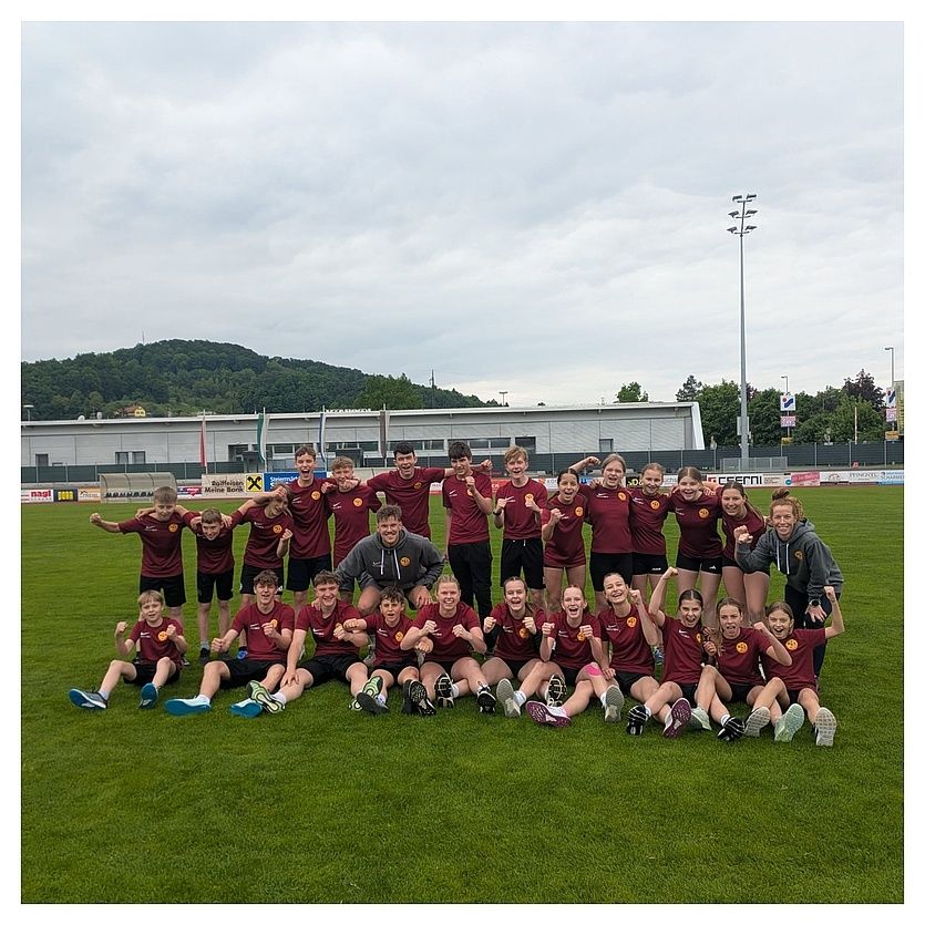 Eine Gruppe junger Athleten posiert für ein Mannschaftsfoto auf einem Feld mit bewölktem Himmel, Bergen und einem Gebäude im Hintergrund.
