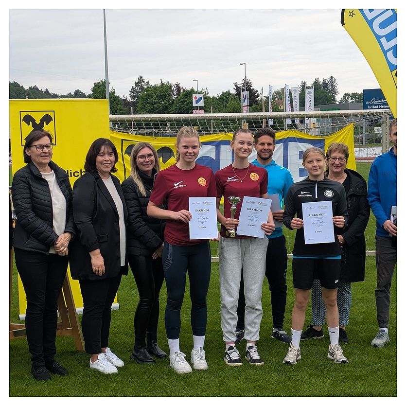 Eine Gruppe von Menschen steht auf einem Fußballfeld mit Trophäen und Zertifikaten, lächelt und posiert für ein Foto. Einige halten Zertifikate und Trophäen.
