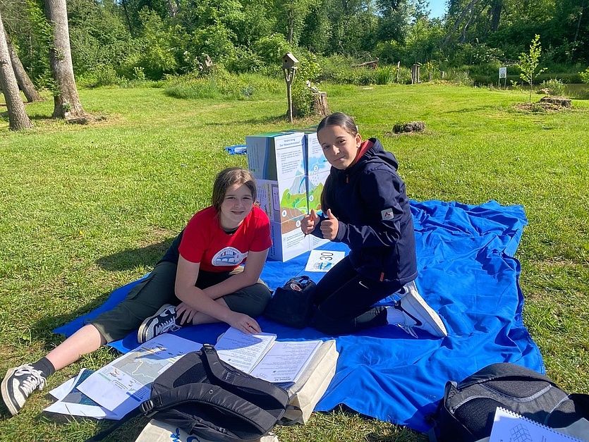 Zwei junge Frauen sitzen auf einer blauen Decke in einem Park, eine gibt Daumen hoch. Sie sind umgeben von Papieren, einem Rucksack und einer Aufbewahrungsbox. Bäume und ein Vogelhaus sind im Hintergrund.