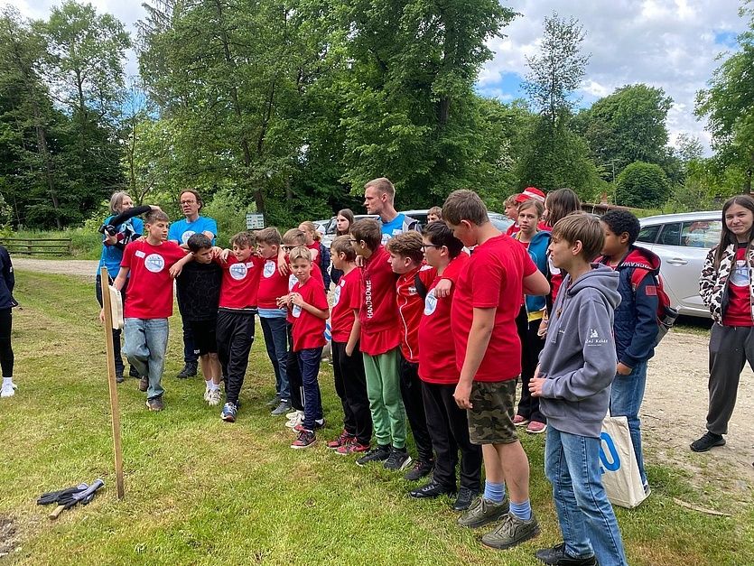 Eine Gruppe von Kindern in roten T-Shirts, wahrscheinlich bei einer Schulveranstaltung, steht auf einem Rasenfeld. In der Ferne hält eine Person eine Kamera.