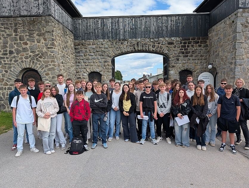 Eine Gruppe von Schülern steht vor einem steinernen Torbogen und lächelt für ein Foto. Einige tragen Sonnenbrillen, Rucksäcke und Sneakers. Hinter ihnen ist ein Schild an der Wand angebracht.