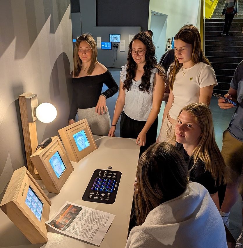 Eine Gruppe von Frauen ist um einen Tisch mit Bildschirmen versammelt, möglicherweise in einer interaktiven Ausstellung. Eine Person sitzt, und eine andere steht hinter dem Tisch.