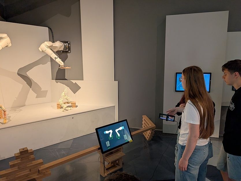 Eine Gruppe von Menschen befindet sich in einem Raum mit einem großen Monitor, einem mechanischen Arm und einer Holzstruktur. Die Menschen schauen auf den Monitor und scheinen an einer robotergesteuerten Aktivität beteiligt zu sein.