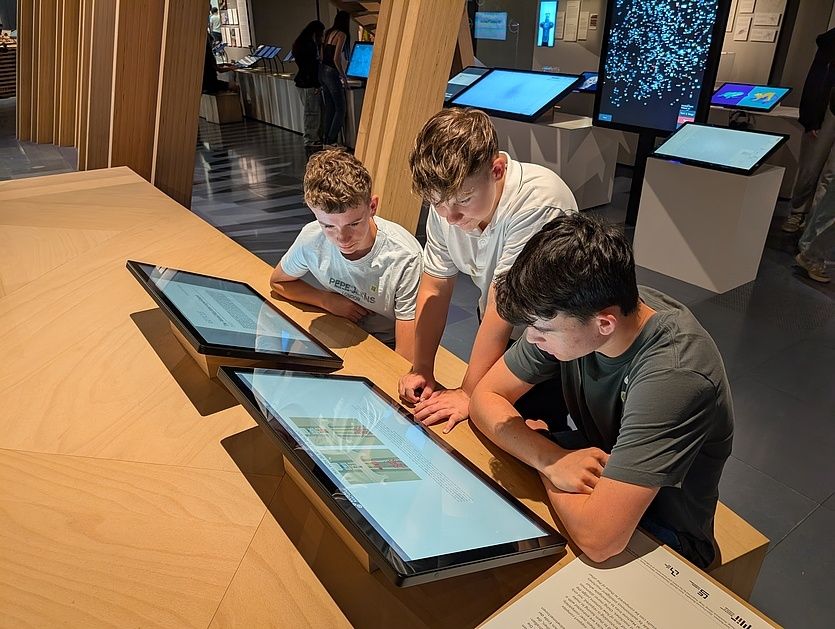 Drei Jungen nutzen ein Touchscreen-Tablet in einem Museum. Sie sitzen um einen Tisch herum mit drei Tablets. Ein weiterer Junge steht hinter ihnen.