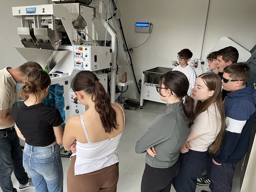 Eine Gruppe von Schülern ist um eine große Maschine versammelt, wahrscheinlich in einem Labor, beobachtet und diskutiert. Die Maschine hat zwei Zahlen, 1 und 2, auf ihrer Vorderseite.