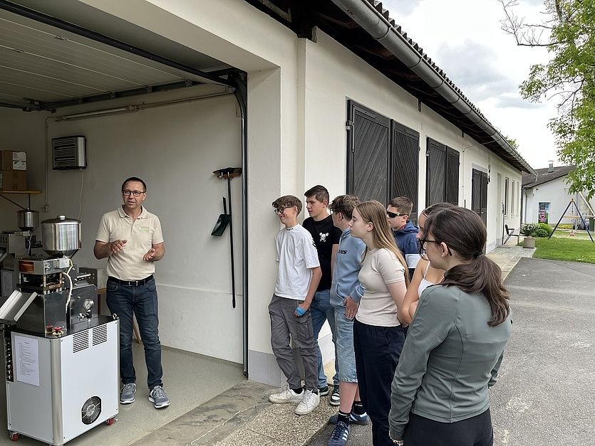 Eine Gruppe von Schülern steht vor einem Gebäude und hört einem Mann zu, der etwas erklärt. Der Mann steht vor einer Maschine.