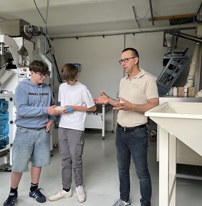 Drei Personen stehen in einer Fabrik. Zwei Jungen halten eine Schüssel und eine Röhre. Der Mann spricht mit ihnen. Industriemaschinen sind hinter ihnen.