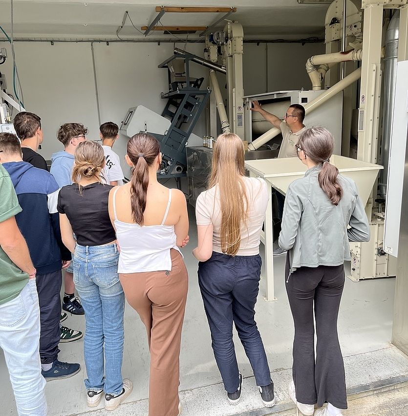 Mehrere Personen versammeln sich in einem Raum mit Industrieanlagen. Ein Mann steht vorne, erklärt etwas und zeigt dabei. Die Leute hören aufmerksam zu.