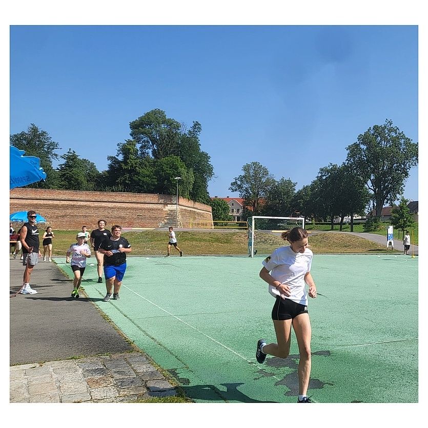 Eine Gruppe von Menschen joggt auf einem grünen Feld. Eine Frau führt die Gruppe an. Im Hintergrund steht ein Fußballtor. Ein Mann steht unter einem Regenschirm.