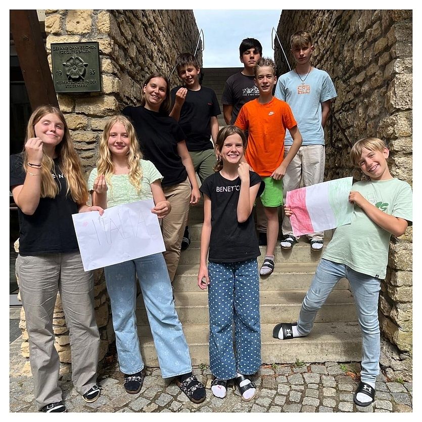 Eine Gruppe junger Menschen, lächelnd und Schilder haltend, steht auf einer Treppe vor einer Steinmauer. Sie tragen Freizeitkleidung und Sneakers.