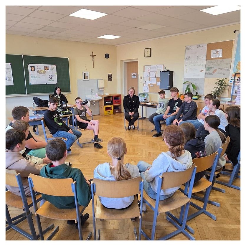 Ein Klassenzimmer mit einer Lehrerin und Schülern. Die Lehrerin sitzt in der Mitte, während die Schüler in einem Kreis um sie herum sitzen. Das Klassenzimmer hat einen Holzboden, eine Pinnwand und ein Waschbecken.