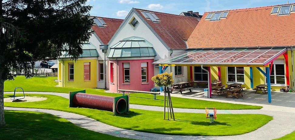 Ein sonniger Tagesausblick auf ein farbenfrohes Kindertagesstätte mit einem Spielplatz, Picknicktischen und Solarpaneelen auf dem Dach. Bäume und Gras umgeben das Gebäude.