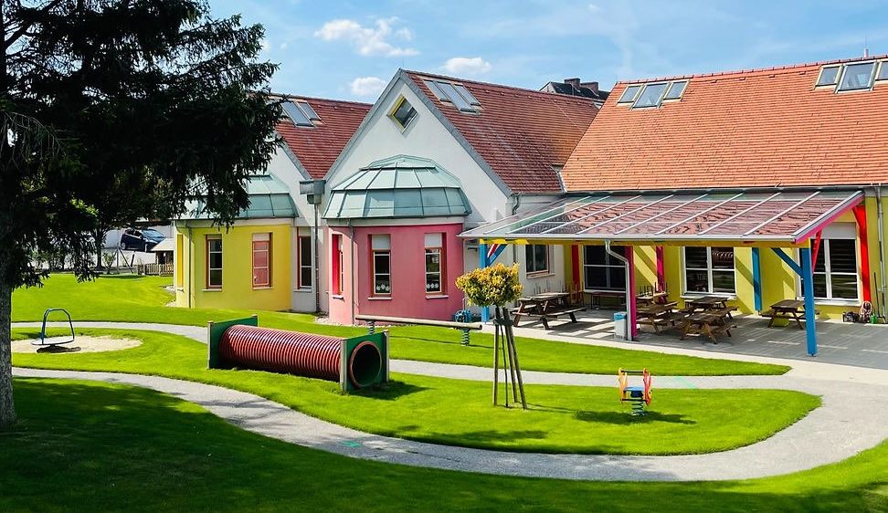 Ein sonniger Tagesausblick auf ein farbenfrohes Kindertagesstätte mit einem Spielplatz, Picknicktischen und Solarpaneelen auf dem Dach. Bäume und Gras umgeben das Gebäude.