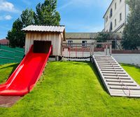 Ein Kinderspielplatz mit einer roten Rutsche und einem Holzhaus. Dahinter steht ein Gebäude mit vielen Fenstern. Bäume und Gras umgeben den Bereich.