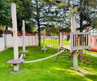 Spielplatz im Freien mit Kletterseilen und Plattformen. Grüner Rasen, Holzpfosten und eine rote Leiter. Bäume und eine weiße Wand im Hintergrund.