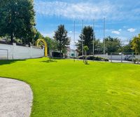 Ein breiter, saftig grüner Rasen mit einem Kiesweg auf der linken Seite, einem gelben Bogen und Bäumen. Ein weißer Wall und ein Metalltor sind im Hintergrund.