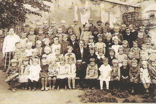 Ein altes Foto zeigt eine große Gruppe von Kindern und einigen Erwachsenen, die vor einem Gebäude stehen und sitzen.