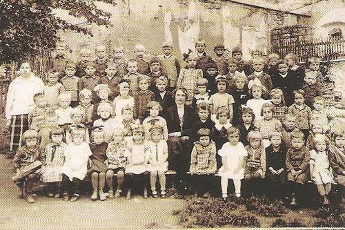 Ein altes Foto einer Gruppe von Kindern und Erwachsenen, wahrscheinlich eine Schulklasse, posiert im Freien vor einem Gebäude.