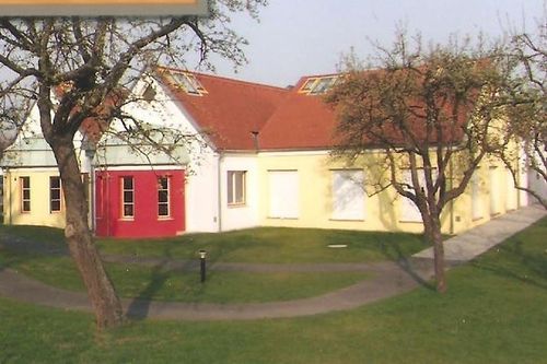 Ein großes gelbes Gebäude mit einem roten Teil hat ein rotes Dach und mehrere Fenster. Es ist von einem grünen Rasen und Bäumen umgeben.