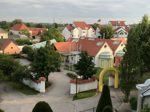Ein Blick von oben auf eine europäische Stadt mit terrakottafarbenen Dächern, umgeben von üppigem Grün und Bäumen. Ein gelber Bogen markiert den Eingang.