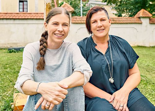 Zwei Frauen sitzen auf einer Bank, beide lächeln, in einer Außenumgebung mit einer Wand und Bäumen im Hintergrund.