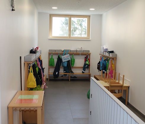 Ein sauberer Raum mit weißen Wänden und Boden hat Holzbänke und Haken mit verschiedenen Taschen und Jacken, die daran hängen. Der Raum hat ein großes Fenster mit Bäumen draußen.