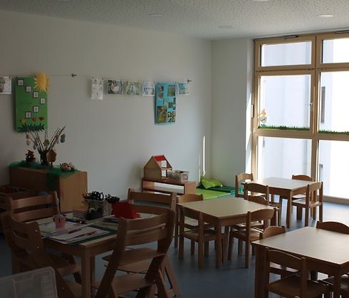 Ein Klassenzimmer mit Holztischen und Stühlen, Wänden, die mit Bildern und einer grünen Tafel dekoriert sind, großen Fenstern rechts und einem Schrank links.