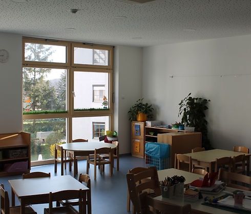 Ein Klassenzimmer mit großen Fenstern, Holztischen und Stühlen, einem Regal mit Pflanzen und einer Uhr an der Wand. Der Raum ist gut beleuchtet mit Sonnenlicht.