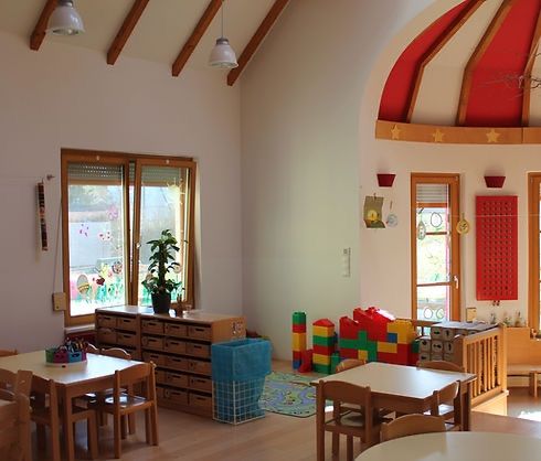Ein gut beleuchteter Spielraum mit Holzmöbeln, einschließlich Tischen und Stühlen, einem Holzschrank, einer Topfpflanze und verschiedenen Spielzeugen. Es gibt Fenster mit Jalousien und dekorative Elemente an der Wand.