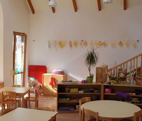 Ein Raum mit Holzboden und weißen Wänden. Es gibt Tische und Stühle herum. Eine Pflanze in einem grünen Topf steht auf einem Regal. Ein Heizkörper ist an der Wand in der Nähe der Tür.