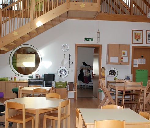 Ein Klassenzimmer mit Holztischen und Stühlen, einer Treppe und einem runden Spiegel. Eine Uhr und Bilderrahmen schmücken die Wände. Schränke und ein Mülleimer befinden sich in der Nähe der Wand.