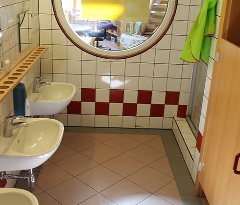 Ein Badezimmer mit gefliesten Wänden und einem runden Spiegel. Zwei weiße Waschbecken und ein grünes Handtuch sind zu sehen. Der Boden ist mit Fliesen bedeckt.