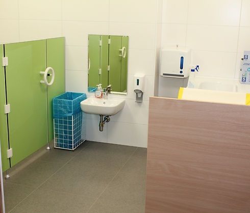 Ein Badezimmer mit einem Waschbecken, einem Spiegel und einem Spender an der Wand. Grüne Trennwände und ein blauer Abfalleimer sind ebenfalls vorhanden.