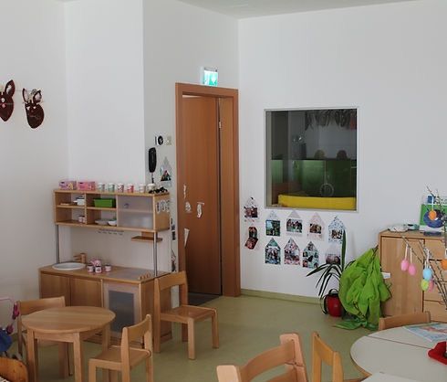 Ein Spielzimmer mit weißen Wänden, einer Holztür, einem Regal und Tischen und Stühlen. An der Wand befinden sich Bilder und Dekorationen, einschließlich einer Topfpflanze.
