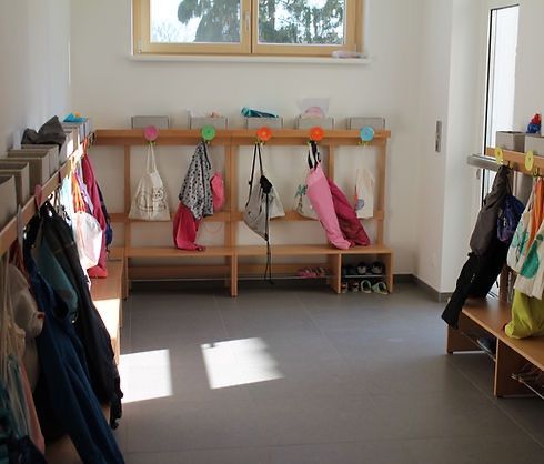 Ein leerer Raum mit weißer Wand und Boden. An der Wand montierte Holzregale mit Taschen und Schuhen, die daran hängen. Das Sonnenlicht kommt durch das Fenster herein.