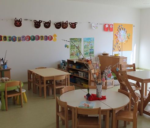 Ein Kindergartenzimmer mit runden Tischen und Stühlen. Die Wände sind mit bunten Dekorationen und Katzengesichtern verziert. Eine Vase mit Blumen und ein Buch stehen auf dem Tisch.