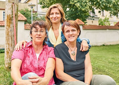 Drei Frauen sitzen lächelnd auf Gras. Die Frau in Pink trägt eine Brille. Die Frau in Gelb hat lange blonde Haare. Die Frau in Schwarz trägt eine Halskette.