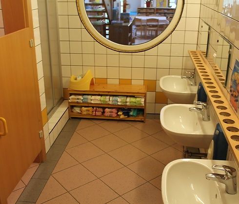 Ein kleines Badezimmer mit gefliesten Wänden, drei Waschbecken und einem runden Spiegel, der einen Raum mit Tisch und Stühlen reflektiert.