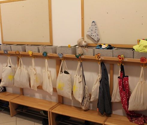 Ein Raum mit weißen Wänden, Holzregalen und weißen Taschen mit gelben Sonnen. Es gibt Aufbewahrungsboxen, Spielzeug und Kleidung auf den Regalen. Zwei Whiteboards sind an der Wand montiert.