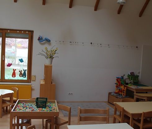 Ein Raum mit Holzmöbeln und einer weißen Wand, einem Fenster mit Außenblick, einem kleinen Tisch mit bunten Kugeln, einer Vase mit Blumen und einem Heizkörper an der Wand.