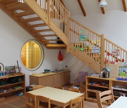 Eine hölzerne Treppe führt in einen hellen, kinderfreundlichen Raum mit Waschbecken, Spiegel, Regalen und einem Tisch. Die Wände sind weiß und der Raum ist durch natürliches Licht erhellt.