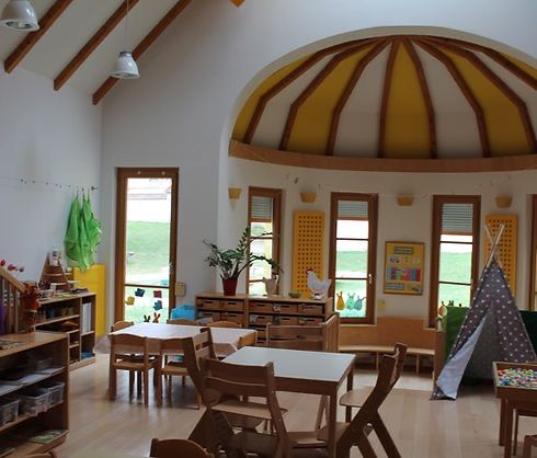 Ein gemütliches Klassenzimmer mit Holzmöbeln, einer runden Decke und Fenstern, die einen Blick auf ein Grasfeld draußen bieten. Es gibt einen Tisch mit Stühlen, eine Bank und ein Tipi.