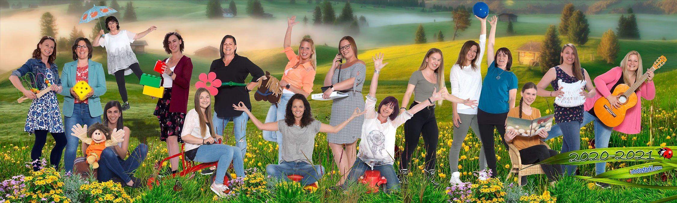 Eine Gruppe von Frauen in Freizeitkleidung steht in einem Feld voller gelber Blumen, einige knien, lächeln und winken. Sie sind in verschiedenen Posen, und einige halten Requisiten. Der Hintergrund zeigt eine neblige Landschaft mit Bäumen und einem Haus.