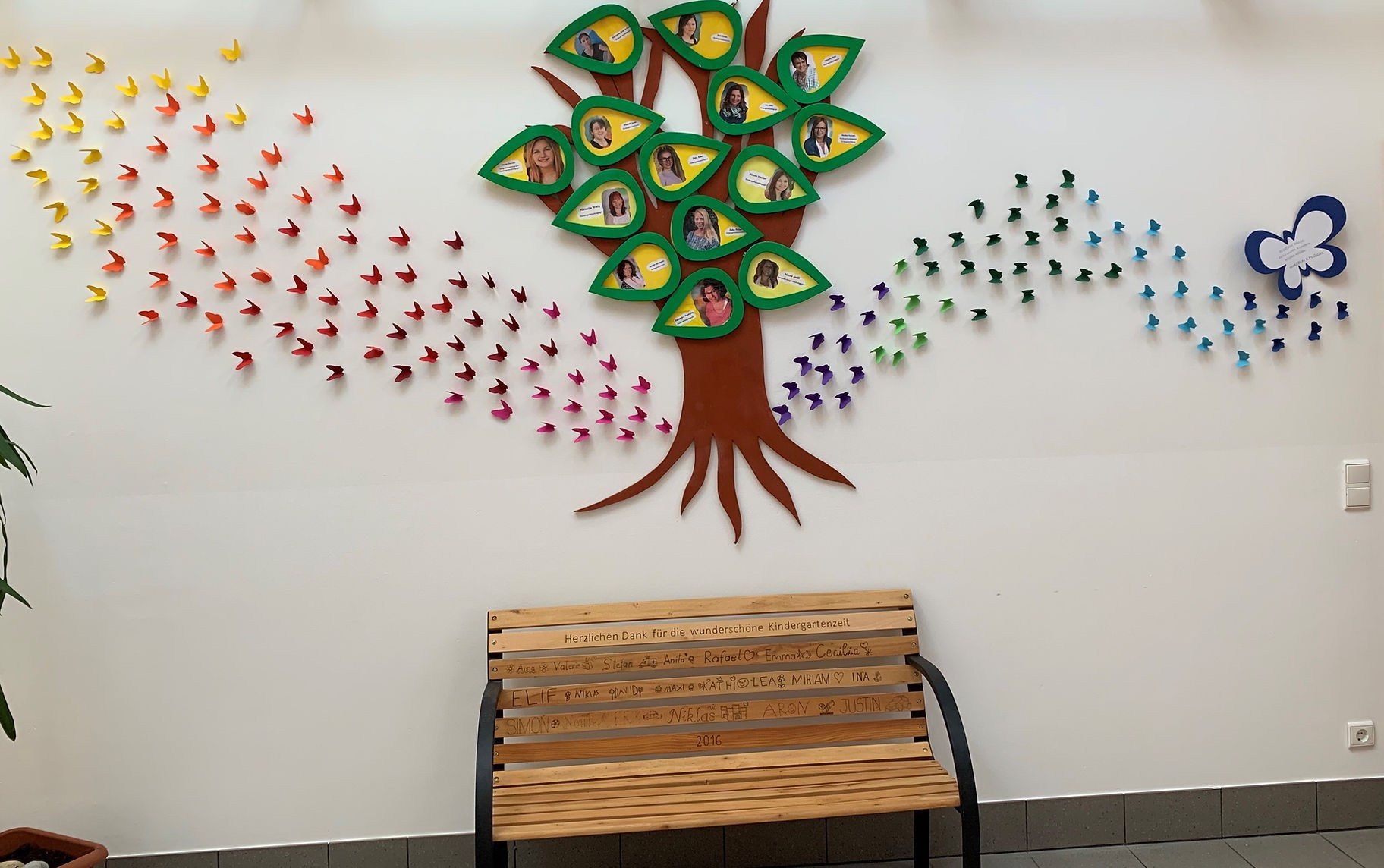 Ein Holzbank mit eingravierten Namen steht vor einer Wand mit einem Baum aus Fotos. Der Baum hat grüne Blätter mit Fotos von Kindern und Erwachsenen. Die Wand hat eine bunte Schmetterlingsdekoration.