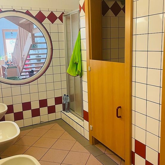 Ein Badezimmer mit gefliesten Wänden, zwei Waschbecken und einer Tür. Ein Spiegel reflektiert eine Treppe und eine Pflanze.
