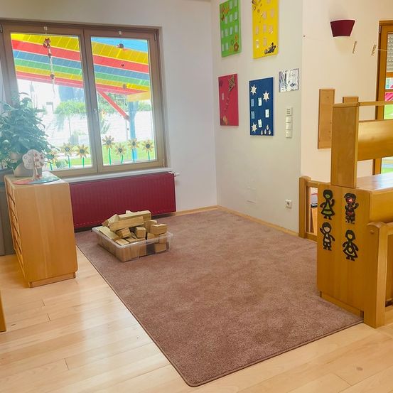 Ein gemütliches Zimmer mit Holzmöbeln und einem Teppich, einem Fenster mit bunten Kunstwerken, einem hölzernen Schrank mit einer Pflanze und einem Heizkörper.