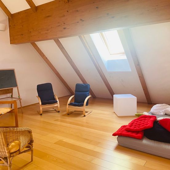 Ein Dachzimmer mit Holzboden, zwei blauen Stühlen, einer Tafel, einem weißen Hocker und einem Bett mit Decken. Es gibt eine Wandlampe und ein Dachfenster.