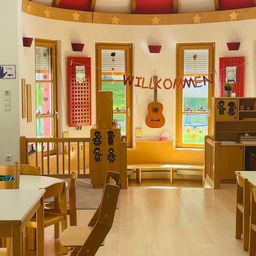 Ein sonniger Raum mit einer Holzbank, einem Tisch und Stühlen. Der Raum hat eine Gitarre, Regale mit Büchern und ein Banner mit der Aufschrift 'Willkommen'. Die Wände sind mit Sternen verziert, und es gibt drei Fenster mit roten Rolläden.