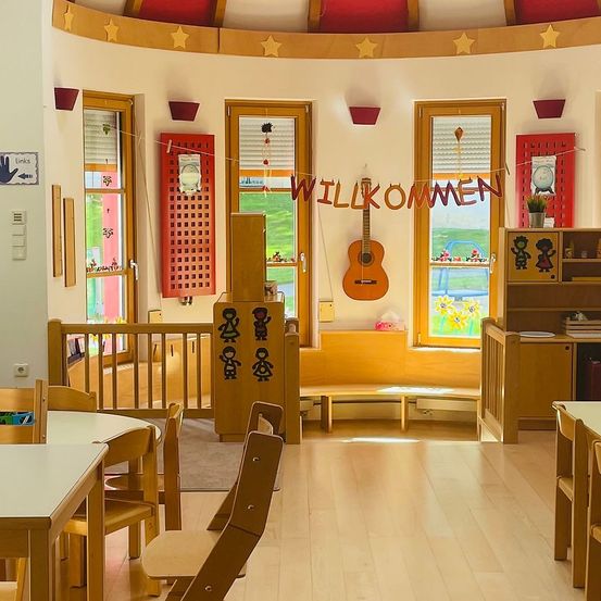 Ein sonniger Raum mit einer Holzbank, einem Tisch und Stühlen. Der Raum hat eine Gitarre, Regale mit Büchern und ein Banner mit der Aufschrift 'Willkommen'. Die Wände sind mit Sternen verziert, und es gibt drei Fenster mit roten Rolläden.