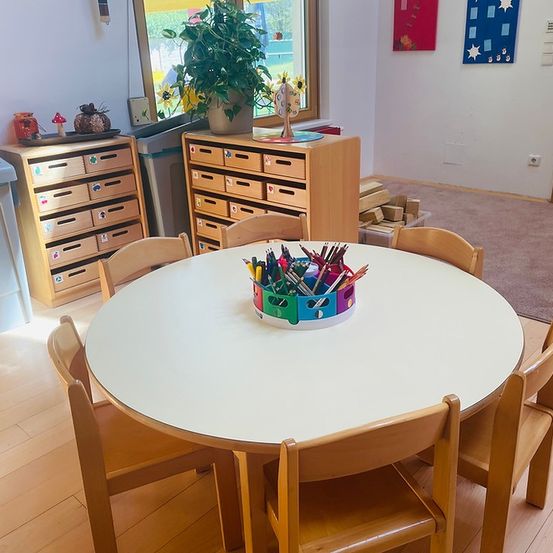 Ein Klassenzimmer mit Holzmöbeln, einem runden Tisch mit einem Korb bunter Stifte, einem Topf mit Pflanzen und einer Wand mit bunten Dekorationen.